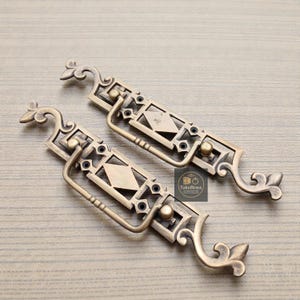 Peut inclure: Deux poignées de tiroir en laiton antique avec des motifs géométriques ornés. Chaque poignée présente une plaque arrière rectangulaire avec un centre en forme de losange et des accents décoratifs. Les poignées ont une poignée incurvée et des extrémités décoratives. Le logo "TukuBrass" est visible.