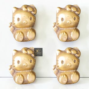 May include: Four brass-colored cabinet knobs in the shape of a cartoon cat. Each knob is detailed with a bow, facial features, and seated pose. The knobs are arranged in a square pattern against a white background.