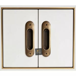 May include: Close-up of a white cabinet with two recessed brass-colored handles. The cabinet doors have a rectangular shape with a gold trim. A small tag with text is visible between the handles.