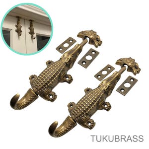 以下が含まれることがあります： ブロンズ色のワニの形をしたドアラッチ2つと、それに合う取り付けプレート。ラッチはフックと詳細なワニのデザインが特徴です。画像には、ドアフレームに取り付けられた2つのワニのフックも表示されています。