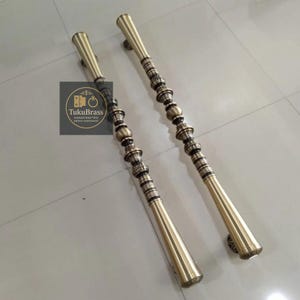 May include: Two brass door handles with ornate detailing. The handles have a long, cylindrical shape with decorative elements and a flared base. The handles are gold in color and appear to be made of metal. The logo "TukuBrass" is visible.