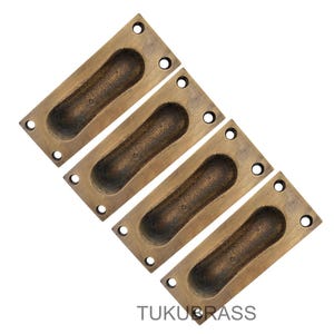 May include: Four antique brass recessed pulls. Each rectangular pull has a rounded, oval-shaped finger pull and four screw holes. The pulls are a warm, aged brass colour with darker accents in the recessed areas. The text "TUKUBRASS" is at the bottom.