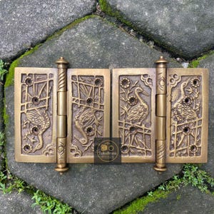 May include: Ornate brass door hinges with detailed crane and floral relief carvings. The hinges are a warm, antique gold color and feature a decorative design. The image shows the hinges open, revealing the intricate craftsmanship and the TukuBrass logo.