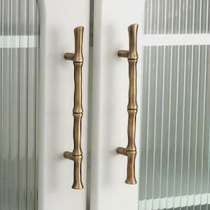 May include: Two bronze-coloured cabinet door handles with a bamboo design. The handles are mounted on a white cabinet door with a glass panel. The handles have a vertical orientation and a textured surface.