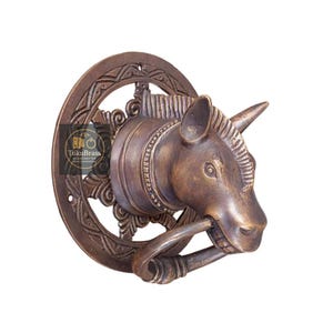 May include: A bronze unicorn head door knocker with a ring in its mouth, mounted on a circular, ornate plate. The knocker is a detailed sculpture with a horn and intricate carvings. The InkuBrass logo is visible on the left side.