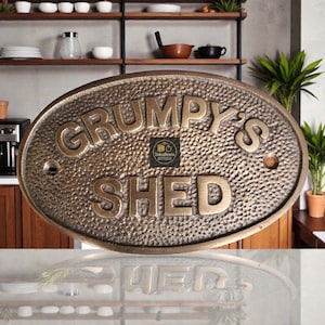 以下が含まれることがあります： 「GRUMPY'S SHED」の文字が浮き彫りになった楕円形のブロンズ看板。表面はテクスチャ加工が施されており、木製の背景に取り付けられています。小屋やワークショップに適しています。
