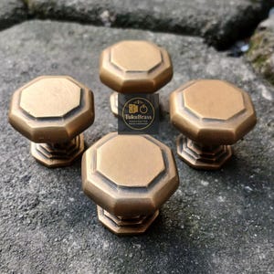 May include: Four antique brass cabinet knobs with an octagonal design. The knobs have a warm, aged finish and are arranged on a textured, grey surface. The knobs are likely for furniture restoration or adding a vintage touch to cabinetry.