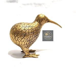 May include: A golden brass kiwi bird figurine. The bird has a long beak and detailed feather texture. The figurine is standing on a white surface. The logo "TukuBrass" is visible.