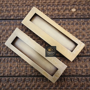 May include: Two rectangular brass cabinet pulls with recessed finger pulls. The pulls are a warm gold colour and are set against a textured brown background. The logo "TukuBrass Handcrafted Hardware" is visible.