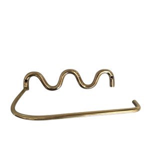 May include: A gold-coloured metal paper towel holder. The design features a wavy top section and a curved bar for holding the paper towels. The holder is designed to be mounted on a wall.
