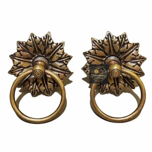 Op de afbeelding: Twee antieke messing deurknoppen. Elke knop heeft een ringhandvat bevestigd aan een decoratief bloemmotief. De knoppen hebben een vintage esthetiek en zijn ideaal om een vleugje elegantie toe te voegen aan elke deur of lade.