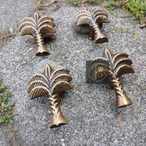 May include: Four bronze-colored palm tree-shaped cabinet pulls. Each pull features a detailed palm frond design and a textured trunk. The pulls are arranged on a gray stone surface.