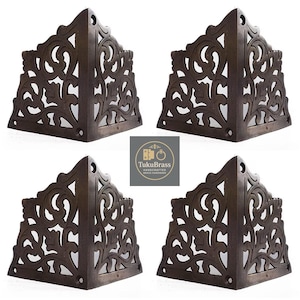 May include: Four decorative, dark brown metal corner brackets with intricate, openwork scroll designs. Each bracket is triangular and features a small hole for mounting. The brackets are arranged on a white background.
