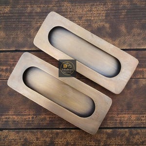 May include: Two antique brass recessed pulls with a rectangular shape and an oval finger pull. The pulls are set against a dark wood background. The logo "TukuBrass" is visible.