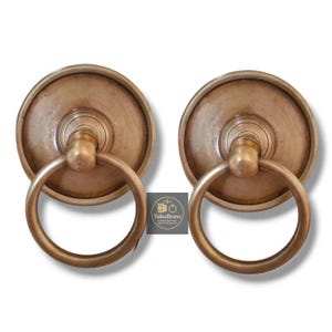 Puede incluir: Dos aldabas de latón antiguo con tiradores de anillo. Cada aldaba tiene una placa trasera circular y un tirador de anillo redondeado. El latón tiene una pátina cálida y envejecida. El logotipo "TukuBrass" es visible.