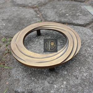 Puede incluir: Un soporte decorativo circular de color latón con un diseño en espiral. El soporte tiene tres patas cortas y una abertura central. La superficie tiene un aspecto texturizado. El logotipo "TukuBrass" es visible.