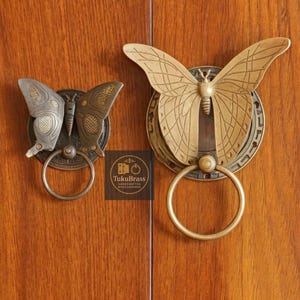 Könnte beinhalten: Zwei schmetterlingsförmige Schrankgriffe in Messing- und Bronzetönen. Die Griffe weisen komplizierte Flügeldarstellungen und einen kreisförmigen Ringgriff auf. Das Bild enthält den Text "TukuBrass HANDCRAFTED BRASS HARDWARE".