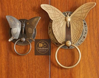 Puede incluir: Dos tiradores de armario con forma de mariposa en tonos latón y bronce. Los tiradores presentan intrincados detalles de alas y un tirador de anillo circular. La imagen incluye el texto "TukuBrass HANDCRAFTED BRASS HARDWARE".