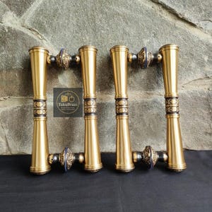 May include: Four brass door handles with a gold finish and intricate black and blue detailing. The handles have a cylindrical shape with decorative elements. The image includes a logo that reads "TukuBrass Handcrafted Door Handles".