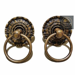 May include: Two antique brass door pulls. Each pull features a circular backplate with an ornate floral design and a round pull ring. The hardware has a vintage aesthetic, with a dark bronze finish and a classic design.