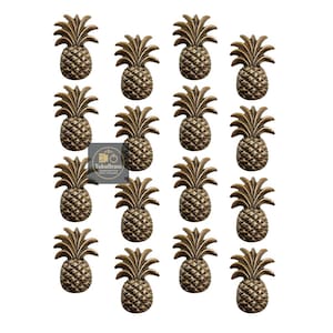 May include: A collection of bronze-colored pineapple-shaped cabinet knobs. Each knob features detailed textures and a realistic pineapple design. The knobs are arranged in a grid pattern against a white background. The image includes the text "TukuBrass" on a small label.