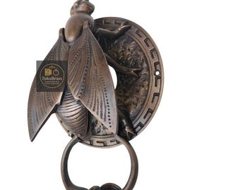 May include: Bronze-colored door knocker in the shape of a detailed insect with large wings and a circular handle. The knocker is mounted on a decorative circular backplate. The brand name "TukuBrass" is visible.