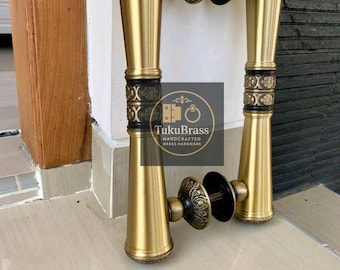 Könnte beinhalten: Ein Paar Messing-Türgriffe mit goldener Oberfläche. Die Griffe haben ein dekoratives schwarzes Band und verzierte runde Enden. Der Text "TukuBrass HANDCRAFTED BRASS HARDWARE" ist sichtbar.
