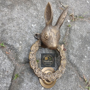 Puede incluir: Un aldaba de puerta de color bronce con forma de cabeza de conejo y orejas largas. El anillo de la aldaba está adornado con diseños de rosas. El texto "TukuBrass HANDCRAFTED BRASSWARE" es visible.