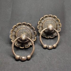 May include: Two antique bronze-colored cabinet pulls. Each pull features a sunflower-shaped backplate with a ring handle. The pulls have a vintage aesthetic, perfect for adding a touch of elegance to furniture.