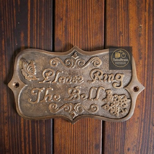 May include: Bronze door sign with the words "Please Ring The Bell" in ornate script. The sign features decorative flourishes, a butterfly, and a snowflake design. It is mounted on a wooden door.