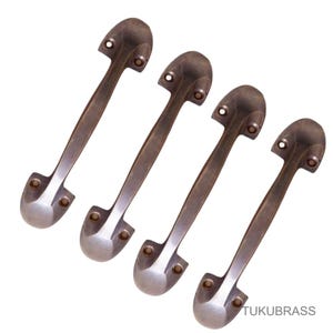 May include: Four bronze door pulls with a classic design. Each pull has a curved handle and rounded ends with mounting holes. The pulls are arranged diagonally against a white background. The brand name "TUKUBRASS" is visible.