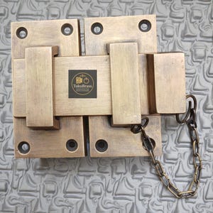 May include: A brass-colored, rectangular door latch with a chain. The latch is composed of several rectangular pieces, with a central square section and a smaller rectangular piece on each side. The latch has the text "TukuBrass" on it.