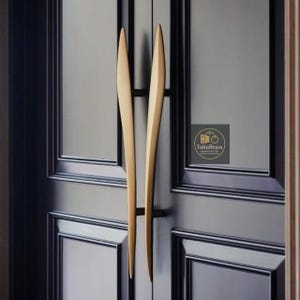 Puede incluir: Primer plano de una puerta negra con dos largos tiradores dorados. Los tiradores tienen un diseño elegante y curvado. La puerta tiene un diseño de paneles rectangulares. El logotipo "TukuBrass HANDCRAFTED" está en la esquina inferior derecha.