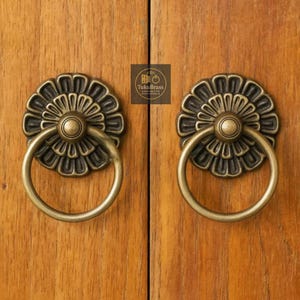 Peut inclure: Deux poignées de meuble en laiton antique avec un motif floral. Chaque poignée présente une plaque arrière circulaire avec un motif de pétales détaillé et une poignée en anneau rond. Les poignées sont montées sur une porte d'armoire en bois. Le logo "TukuBrass" est visible.