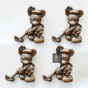 May include: Four bronze-toned Mickey Mouse cabinet pulls. Each pull depicts Mickey Mouse in a seated position, holding a golf club. The pulls have a vintage finish, ideal for adding a touch of fun to furniture.
