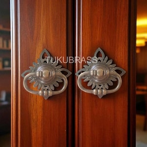 May include: Two antique-style cabinet pulls with a floral design. The pulls are silver-toned with a ring handle and are mounted on a dark wood cabinet. The text "TUKUBRASS" is visible above the pulls.