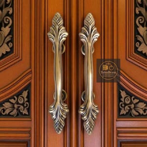 May include: Two ornate brass door handles with a leaf design are mounted on a wooden door. The handles are vertically oriented and have a detailed, textured appearance. The door has decorative carvings.