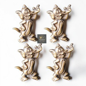 May include: Four brass cat-shaped cabinet pulls. Each pull features a cartoon cat design with outstretched arms and a smiling face. The pulls are in a standing pose and have a vintage, antique appearance. The TukuBrass logo is visible.