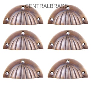 Peut inclure: Six poignées coquilles de style antique, de couleur bronze. Chaque poignée a une forme en demi-cercle avec une série de lignes verticales rayonnant du centre. Le texte "CENTRALBRASS" est imprimé en haut.