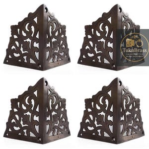 Puede incluir: Cuatro escuadras de esquina de color marrón oscuro con diseños calados ornamentados. Cada escuadra es triangular, con un patrón decorativo de líneas y curvas arremolinadas. Las escuadras están hechas de un material marrón oscuro y sobre un fondo blanco.