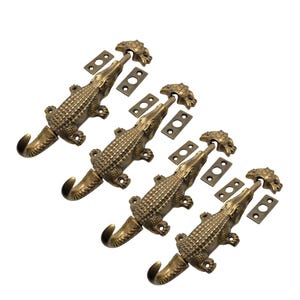 May include: Four antique brass-colored alligator-shaped door latches. Each latch features a hook, a detailed alligator body, and a decorative head piece. Small rectangular plates and screws are included.