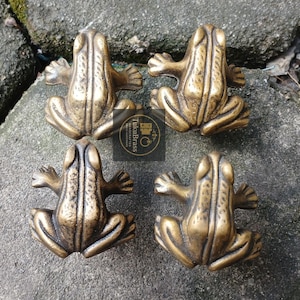 May include: Four bronze frog-shaped cabinet pulls. Each pull features a detailed frog design with outstretched arms and legs. The pulls are arranged on a grey stone surface. The text "TukuBrass HANDCRAFTED" is visible.