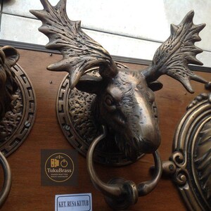 May include: Bronze-toned door knocker featuring a detailed moose head with large antlers. The moose head is mounted on a wooden panel, with a circular design around it. The knocker includes a ring for knocking.