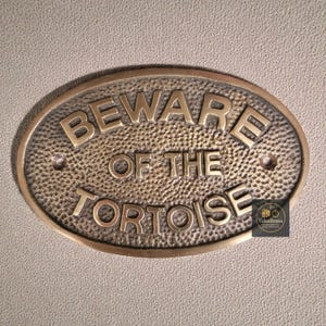 May include: Oval brass plaque with the words "BEWARE OF THE TORTOISE" in raised lettering. The plaque has a textured background and two small holes for mounting. The TukuBrass logo is in the lower right corner.