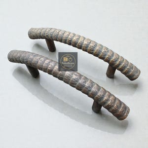 Pode incluir: Duas puxadores de armário curvos de cor bronze com um design texturizado e canelado. Os puxadores são arqueados e têm uma aparência rústica e antiga. Os puxadores são feitos de metal e são adequados para móveis. O logotipo "TukuBrass" é visível.