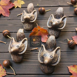 Könnte beinhalten: Vier bronzefarbene Schrankknöpfe in Fuchsform sind auf einer dunklen Holzoberfläche angeordnet, umgeben von Herbstblättern und Eicheln. Die Knöpfe weisen detaillierte Merkmale auf, darunter spitze Ohren und strukturiertes Fell. Das Logo "TukuBrass" ist sichtbar.