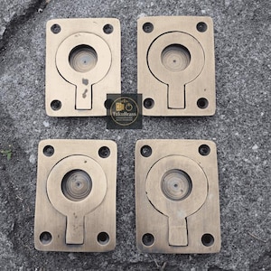 May include: Four antique brass flush pull handles. Each handle is rectangular with a circular recessed pull and a keyhole-shaped cutout. The handles are mounted with four screws. The logo "TukuBrass" is in the center.