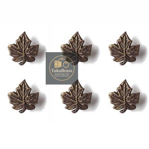 May include: Six bronze-coloured leaf-shaped cabinet knobs. Each knob has detailed veining and a slightly raised texture. The knobs are arranged in two rows of three. The TukuBrass logo is visible.