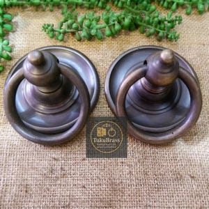Pode incluir: Duas puxadores de armário de bronze antigo com puxadores de anel. Cada puxador tem uma placa traseira circular e um botão arredondado. O logotipo "TukuBrass Handcrafted Brass Hardware" é visível.