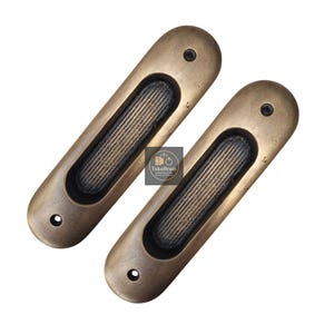 May include: Two antique brass-coloured recessed pulls. Each pull has a long, oval shape with a textured, dark brown centre and two small holes. The pulls are designed for a flush mount, suitable for cabinets or drawers.